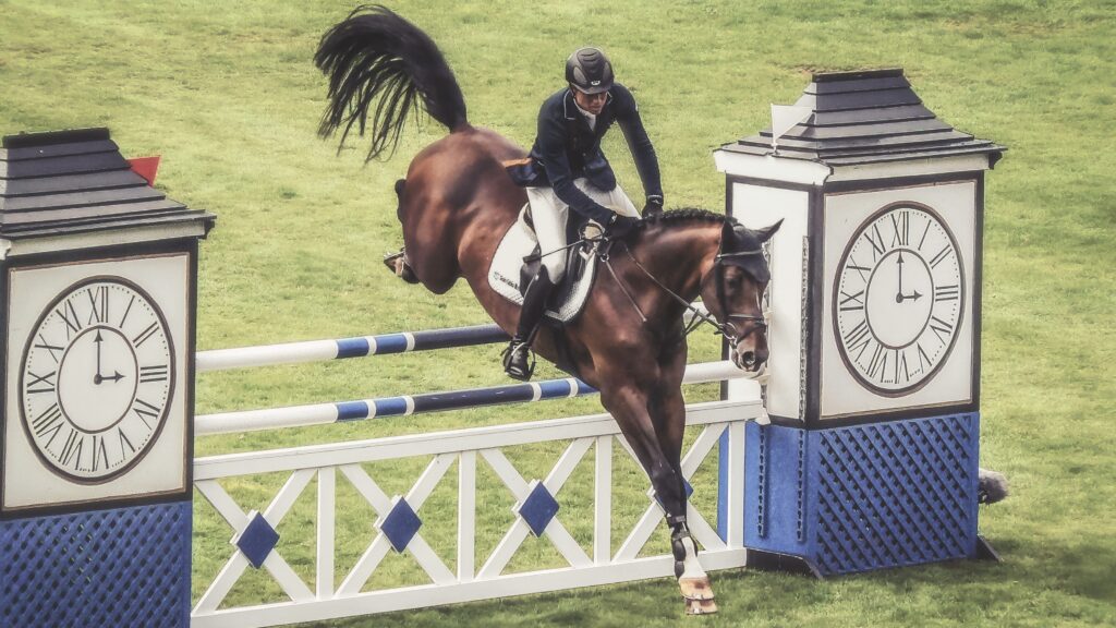 Show jumper riding a bay horse over a fence between two clock-tower standards on a grassy arena.